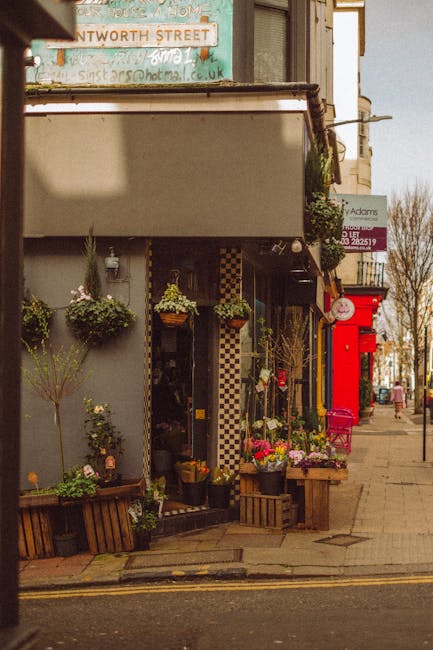 A display of potted flowering plants with vibrant purple, pink, and white blooms arranged on a black metal shelf inside a glass-fronted garden shop. The pots are metallic silver and black, and the dis